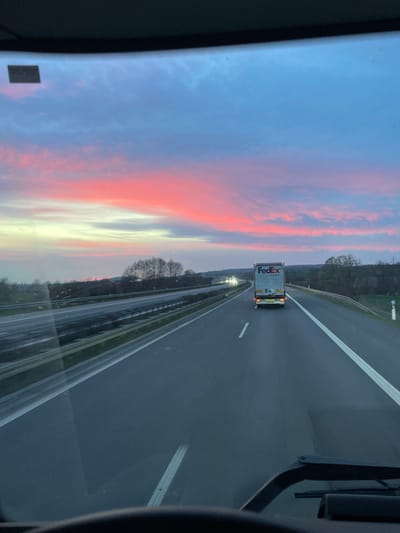 The image depicts a highway scene at twilight, from the interior perspective of a moving vehicle, looking forward through the windshield. The location is Wallhausen, Germany. The sky above is a prominent feature, displaying a gradient from deep blue at the top, transitioning through lighter blue and purple, to a vibrant horizontal band of intense pink and red clouds across the mid-horizon. Below this, the sky lightens to a pale yellow-white.

The foreground shows the upper portion of a vehicle's dashboard and a segment of a steering wheel. The multi-lane asphalt highway appears dark, with distinct white solid and dashed lane markings. Ahead, a white semi-trailer truck with "FedEx" branding (purple and orange logo) is visible in the same lane, its taillights illuminated. To the left, a median barrier separates the vehicle's lanes from opposing traffic lanes, where faint headlights from other distant vehicles are discernible. The roadside features bare trees and general vegetation, indicating a non-foliage season. Undulating terrain or hills are visible on the horizon beneath the vibrant sky. White roadside reflector posts are present along the right shoulder.