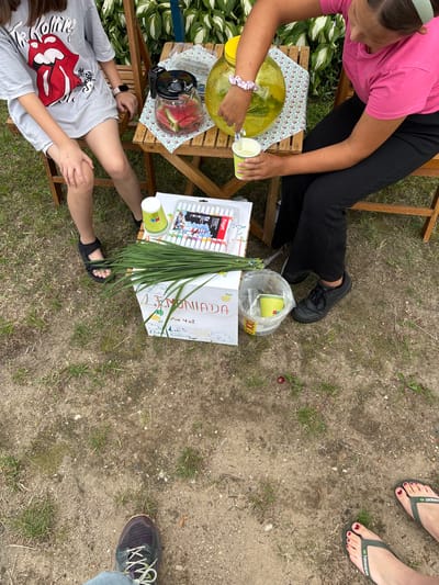 In Trześcianka, Poland, two young females are seated on wooden chairs at a wooden table outdoors. One female, wearing a grey t-shirt with a Rolling Stones logo, sits on the left. The other female, in a pink t-shirt and black pants, is on the right, pouring a clear yellow liquid from a large glass dispenser into a small green paper cup. On the table are a glass pitcher filled with watermelon slices, a floral-patterned cloth, and a box of markers. A box labeled "Lemoniada" with drawings is on the ground next to a bunch of long green grass. The ground is covered with dirt and sparse grass. In the lower portion of the image, the feet of two individuals wearing sneakers and flip-flops respectively are visible. The scene is set in a natural, grassy environment.