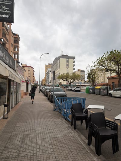 This image depicts a street scene in Grau I Platja, Spain, under an overcast sky. A tiled pedestrian sidewalk occupies the left foreground, where a solitary individual, dressed in dark attire, walks away from the camera. To the right of the sidewalk, multiple stacks of dark plastic chairs and two white rectangular tables are positioned near the curb, partially enclosed by a collapsed blue temporary barrier.
Along the street, a continuous line of parked vehicles, predominantly dark-colored, faces away from the camera. The street features a blue-painted lane and a series of green, yellow, and grey waste bins on its right side. On the left, a row of commercial establishments with awnings includes a storefront partially displaying "Encheguito" and an overhead sign advertising "Bar Cafeteria Restaurante" and "Tapas Manchegas."
In the background, multi-story residential and commercial buildings of various architectural designs and colors (beige, brown, white, grey) line both sides of the street. Several trees with spring foliage are visible throughout the scene, interspersed among the buildings and street furniture. A tall street lamp stands prominently in the midground.
