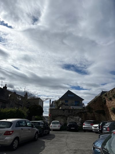 The image depicts a street in Dinard, France, located at latitude 48.63 and longitude -2.053. The scene is a narrow street lined with buildings on either side, with a few cars parked along the street. The sky is partly cloudy, with wispy clouds scattered across the blue sky. The buildings are a mix of residential and commercial properties. The street is paved with asphalt.  There are several cars parked on the street, including a silver hatchback, a white hatchback, a black hatchback, and a blue hatchback. The street is quiet and appears to be residential, with trees lining the street and green foliage visible on the properties.  There is a sign on the wall with a red circle and a diagonal line through it.