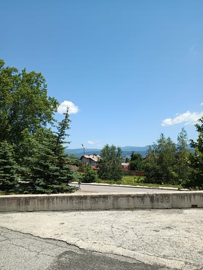 The image shows a view of the city of Zlatitsa, Bulgaria, on a clear day with a bright blue sky and a few scattered clouds. In the foreground, there is a weathered concrete barrier. Beyond the barrier, a road is visible with a lone person walking on the sidewalk. Behind the road, a variety of trees, including evergreen and deciduous varieties, form a lush green backdrop. In the middle ground, residential buildings with red-tiled roofs are nestled among the trees. The background features rolling hills and distant mountains, indicating a semi-rural or suburban setting within Zlatitsa. A lamppost is also present near the houses. The overall impression is one of a peaceful, sunny day in a residential area with natural surroundings.