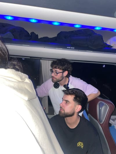 Two young men are in a vehicle. One is wearing glasses and a headset around his neck, with a pink and white striped shirt. The other, closer to the camera, sports a black shirt with a gold emblem. They are looking down. A bus seat is visible in the background. A blue light strip is located next to a window. The scene is slightly rotated.