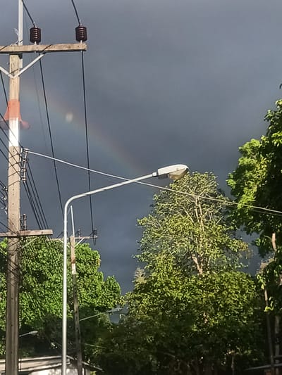 The image captures a partially obscured rainbow arcing across a grey sky, likely after or during a rain shower. In the foreground, there is a utility pole with attached wires and hardware. A street lamp is centered in the image. Lush green trees fill the right side and partially obscure the lower left. The image was taken in Moo 4, Thailand. There are no people visible.