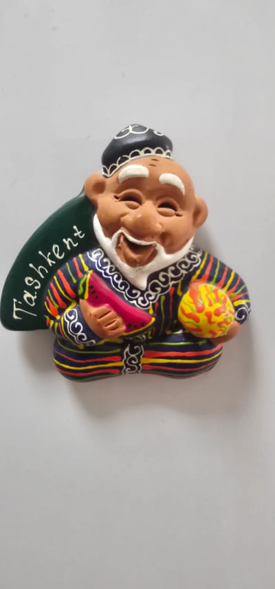 The image shows a ceramic magnet shaped like a jovial old man with a white beard and a traditional cap. He is depicted wearing a colorful striped robe and holding a slice of watermelon in his left hand and a yellow and red ball in his right. The word "Tashkent" is written in white script on a green background to the left of the figure. The magnet is attached to a plain light gray surface, possibly a refrigerator. The specific location of Karshi City, Uzbekistan, is not depicted in the image.