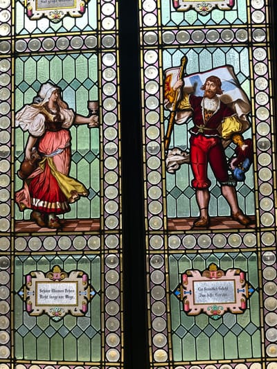 The image shows two stained glass windows. The upper window depicts a man in medieval clothing, holding a flag and a staff. He wears red pants, a maroon jacket, and a white shirt. The flag has a blue and white design. The bottom window features a woman in similar attire, holding a hammer. She wears a red and white dress with a yellow sash. The background of both windows consists of geometric patterns in green and gold. Adjacent to each figure is a framed inscription in German. These stained glass windows are not located in Tlalnepantla, Mexico.
