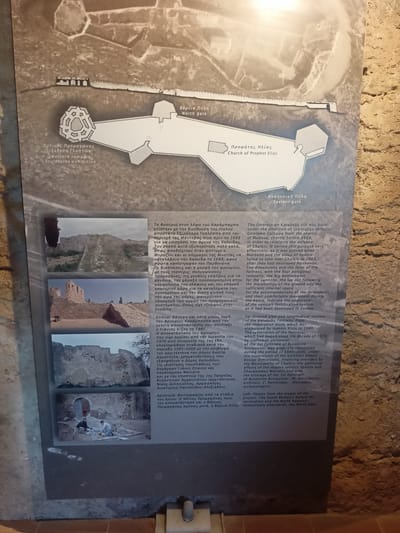 The image shows an informational display board about the Karababa Fortress, a historical site located in Chalkis, Municipal Unit of Anthidon, Greece. The display features a schematic map of the fortress with labels in both Greek and English, indicating points of interest such as the "North Gate" and the "Church of Prophet Elias." Below the map are several photographs depicting different sections of the fortress, including remnants of stone walls and excavation work. The text on the display board provides historical context about the fortress's construction and restoration, mentioning its Italian defectors, Morosini and the Allies of Venice, and its eventual takeover by the Ottomans. It also details the restoration efforts undertaken between 1985 and 2000, highlighting the involvement of the Municipality of Chalkis, the Ephorate of Byzantine Antiquities, and specific architects and archaeologists. The image itself is a photograph of this display board, mounted on a stone wall, with a textured, rough surface typical of ancient structures. The lighting suggests an indoor or shaded outdoor environment, with some light reflecting off the display. There are no people visible in the image of the display board itself, but one of the photographs on the board shows two individuals engaged in archaeological work.