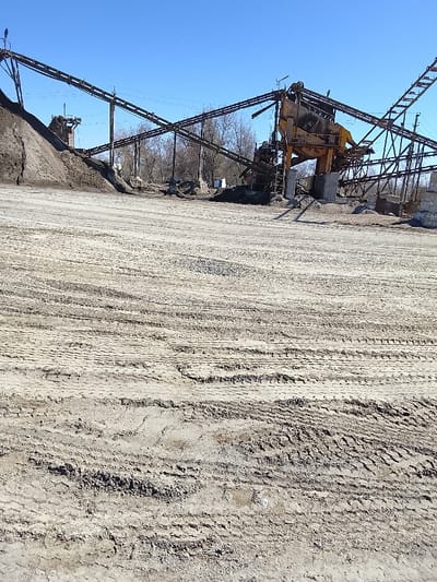 This image depicts an active or disused industrial aggregate processing facility under a clear blue sky in Yangi Chinoz, Uzbekistan. The foreground consists of an expansive, unpaved dirt and gravel ground heavily marked with numerous tire tracks, indicating vehicular traffic. In the midground and background, a complex system of industrial machinery is visible. This includes multiple long, dark-colored conveyor belts supported by lattice metal structures, extending horizontally across the scene. To the left, a large pile of dark brown earth or aggregate rises. To the right, a prominent, large yellow industrial processing unit, possibly a screener or crusher, is centrally located within the conveyor system. Scattered smaller piles of material are visible beneath and around this machinery. Bare deciduous trees are present in the distant background, beyond the industrial equipment. No individuals are visible within the scene, and no overt actions or interactions are depicted. The overall environment suggests an industrial site for material handling.