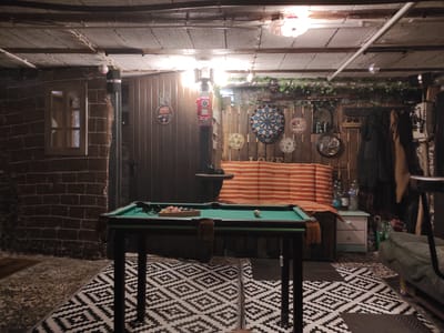 This is a dimly lit, indoor recreation room, possibly a basement or garage, in Waldbronn, Germany. The room features a pool table with billiard balls racked and one ball on the felt. A patterned sofa with orange and white stripes is visible behind the pool table, along with a small white chest of drawers. The walls are constructed of brick and wood paneling, decorated with various circular signs and a dartboard. A red vintage gas pump is mounted on the wall. A window with wooden framing is set into the brick wall. The ceiling is made of concrete tiles and exposed beams, with overhead lighting and some decorative string lights. There is a patterned rug on the floor. A green sofa is partially visible in the lower right corner. Clothing items are hanging on the right side of the room. The overall atmosphere is that of a casual, rustic hangout space.