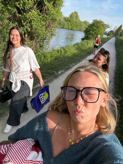 On a sunny day in Marquette-lez-Lille, France, a group of three young women pose for a selfie along a path beside a canal.
The foreground is dominated by a blonde woman wearing eyeglasses, a blue sweater, and a layered gold necklace. She is making a kissing face toward the camera. Behind her and to the left, a brunette woman with sunglasses in her hair is looking downward. To the lower left of the frame, a third brunette woman in a white t-shirt and dark sweatpants smiles as she holds a blue reusable shopping bag and a small black handbag.
The path is bordered on one side by the canal and lush greenery, and on the other side by more greenery. In the distance, a person wearing a safety vest walks along the path. Trees and possibly some buildings are visible further in the background.