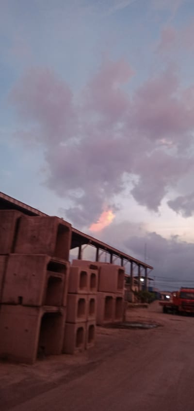 The image shows a scene near Sumput, Indonesia. In the foreground are stacked concrete structures, possibly culverts, suggesting an industrial or construction area. Behind these, a long, open-sided building with a metal roof is visible. In the background, there's a red truck and other structures. The sky is cloudy, with hints of pink and orange near the horizon, indicating either sunrise or sunset. No people are readily visible in the image. The overall environment appears to be a commercial or industrial yard.