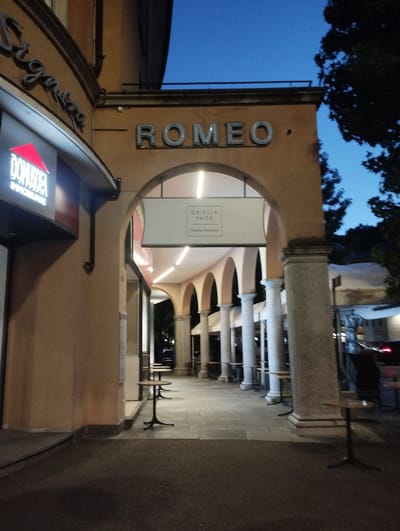 A photograph taken at dusk shows the arched walkway of a building with the word "ROMEO" prominently displayed in large, silver, three-dimensional letters above the arch. To the left, a sign for "DOMUSDEA IMMOBILIARE" is visible. Inside the walkway, several small, round tables with black bases are set up. On the right side of the walkway, a tree with dark foliage is silhouetted against the blue twilight sky. The scene appears to be in Muralto, Switzerland, given the context of the location.