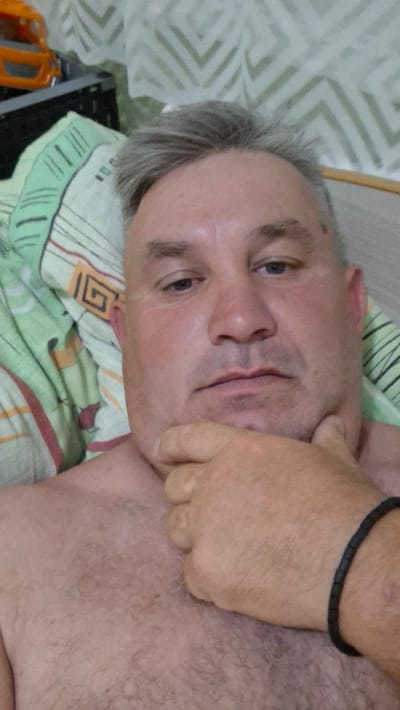 A close-up, waist-up selfie of a light-skinned, middle-aged man with short, graying hair and light blue eyes. He is lying down, with his chin resting on his left hand, which is adorned with a black beaded bracelet. His bare chest is visible. He is wearing a green and beige patterned sheet or blanket. In the background, a wall with a light green zigzag pattern is visible. To the left of the man, part of an orange toy truck can be seen. The scene appears to be indoors, likely in a bedroom, in the city of Voinova, Moldova.