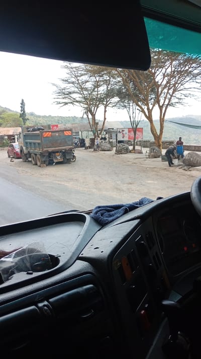 The image, taken from the interior of a vehicle in Lari/Kirenya ward, Kenya, provides a view through the windshield. The foreground displays the vehicle's dark gray dashboard, featuring air vents, control buttons, a gear shifter on the right, and a crumpled blue microfibre cloth resting on the surface. A sun visor is partially visible at the top, and a green-tinted strip is present along the upper edge of the windshield.
Outside the vehicle, a paved road transitions to a dirt shoulder and parking area on the right. A faded blue or green dump truck is parked on the dirt, angled away from the viewer, with its rear bed appearing empty or covered. Reflective strips are visible on its rear. A red sedan is parked further back behind the dump truck. A small motorcycle is parked immediately behind the dump truck's rear.
In the dirt parking area, multiple individuals are present. One person, dressed in dark attire, is bent over, actively working on one of several large, irregular, rounded objects that are painted with black and white stripes resembling zebra patterns. Another individual in a red shirt stands nearby. Other people are visible further back near rudimentary structures with corrugated iron roofs. Numerous similar striped objects are distributed across the area, alongside stacks of tires. A red and white banner, partially obscured, is visible behind the structures; "SAFARICOM" is partially legible on it.
The background features several large trees with light bark and sparse foliage. Beyond these, rolling hills covered with vegetation are visible under a bright, overcast sky.
