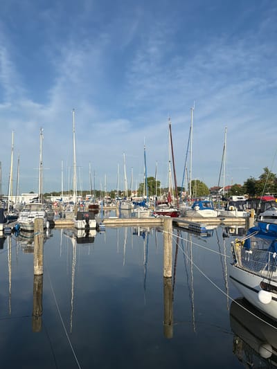 This media file captures a serene marina scene in Ribnitz-Damgarten, Germany. The image showcases a row of sailboats docked along a wooden pier, their masts creating a striking vertical pattern against a clear blue sky with wispy clouds. The water of the marina is calm, reflecting the boats and the sky, adding a sense of depth and tranquility to the composition.
The scene is illuminated by what appears to be the soft light of early morning or late afternoon, indicated by the gentle quality of the light and the lack of harsh shadows. The weather seems pleasant and still, conducive to sailing. The boats are of various sizes and colors, predominantly white with blue and red accents, suggesting a well-maintained fleet. Some boats have their sails furled, while others appear ready for departure.
Notable details include the wooden pilings of the pier, some of which are weathered and bear evidence of their functional purpose in mooring the vessels. The reflections in the water are a significant element, doubling the visual impact of the sailboats and the sky. There are no people visible in the frame, contributing to the peaceful and unoccupied atmosphere.
The location context provided, Ribnitz-Damgarten, Germany, hints at a coastal or lakeside setting known for its maritime activities. The overall mood evoked is one of peace, order, and the quiet beauty of a working marina. There is no visible text in the image.