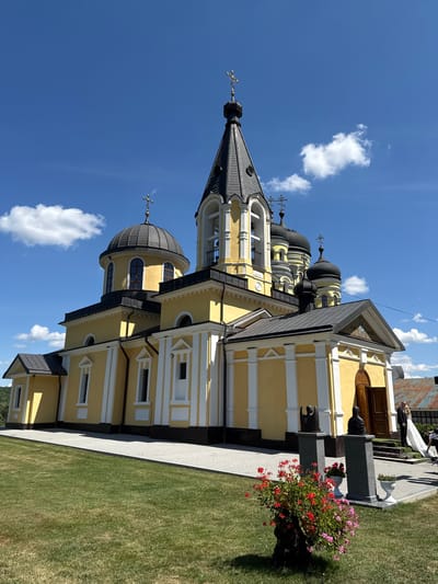 In Ciuciuleni, Moldova, a yellow Orthodox church with multiple dark domes topped by crosses stands under a bright blue sky with scattered white clouds. The church features white decorative columns and trim. In the foreground, a paved walkway leads to the entrance, flanked by green grass. Two dark stone sculptures on pedestals are positioned in front of the church. To the right of the entrance, a man in a dark suit and a woman in a white wedding dress are visible, suggesting a wedding event. A potted plant with red and pink flowers sits in the grassy area to the left of the walkway. The architecture includes bell towers and various rooflines.