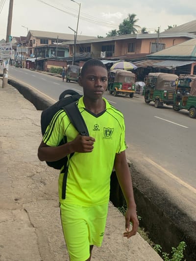 The image depicts a young male standing on a cracked concrete sidewalk in Aba, Abia, Nigeria, facing the camera directly. He is dressed in a neon green short-sleeved collared shirt with black shoulder stripes and trim, featuring a "SPORTS" logo with a soccer ball and a shield emblem. He wears matching neon green shorts with black side stripes and a similar soccer ball logo. A black backpack is on his right shoulder, with his right hand holding the strap.
To the subject's left, a low concrete barrier separates the sidewalk from an asphalt road. Numerous auto-rickshaws (Keke Napep), primarily green and brown/beige, are positioned along the right side of the road, facing away from the viewer. Some vehicles display text such as "Orange" and "4503." Along the left side of the road, a line of multi-story buildings, appearing to be residential or commercial structures with various paint finishes (peach, brown, grey), extends into the distance. In front of these buildings, several ground-level shops with corrugated metal roofs are visible.
Utility poles with overhead lines and streetlights are present along the road. A prominent signboard on a pole on the far left displays political campaign posters, including one with "SOFA 2023." Sparse vegetation is visible in the background and within a concrete drainage ditch adjacent to the sidewalk. The sky is overcast.