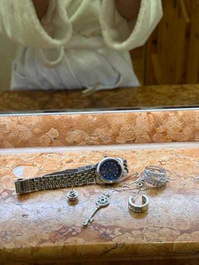 The image captures an indoor scene featuring jewelry laid out on a textured surface, with a reflection of a person and wooden walls on the left.
In the foreground and on the right, various pieces of silver-colored jewelry are arranged on a light brown or tan marble-like surface, possibly a bathroom counter or vanity. These include a watch with a metallic link band and a deep blue face, displaying the text "GENEVE" and "QUARTZ," and "STAINLESS STEEL" on its clasp. Beside the watch, there is a complex, ornate pendant, possibly a pair of intertwined rings, attached to a delicate chain. Also visible are a single hoop earring with ridged detailing, a dangle earring shaped like an elaborate key adorned with small stones, and a single stud earring.
On the left side of the image, a reflection is visible, likely in a mirror or a highly polished surface. This reflection shows a person wearing a white, hooded bathrobe. Only the upper part of their body, including the shoulders and hood, is clearly visible. Behind the person in the reflection, there are wooden panel walls, suggesting a rustic or natural interior setting.
The overall scene suggests a moment of relaxation or preparation in a private accommodation, possibly a hotel or spa, in Going am Wilden Kaiser, Austria, as indicated by the context. The lighting is soft and even, typical of indoor illumination. No specific time of day or weather conditions can be determined.