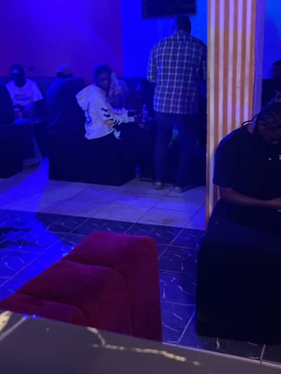 The image depicts an indoor scene, likely a lounge or club, with dim blue lighting. In the foreground, a plush red couch is visible, with a dark, possibly black, seating area to its right. A person in a black t-shirt is seated to the right, looking down at a device. Behind this person, a decorative pillar with vertical light-colored stripes is present. Further back, a person in a red and blue checkered shirt stands facing away from the camera. To the left, two individuals are seated on dark furniture, one in a white hooded sweatshirt and the other in a white t-shirt. The floor appears to be tiled. The overall ambiance is dimly lit and intimate, suggesting a relaxed or social gathering. The context of Durumi, Nigeria is noted, but the visual elements themselves do not provide specific geographical indicators within the city.