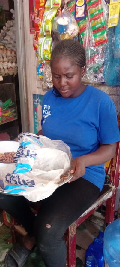 A young woman is seated in what appears to be a small shop in Kwachiri, Nigeria. She is wearing a blue t-shirt with white lettering that reads "FIO PEACE". Her black leggings have a visible hole in the knee. She is holding a white plate containing what looks like peanuts and a white plastic bag with blue and black writing. The woman has dark skin and braided hair.
Behind her, the shop shelves are stocked with various packaged goods, including snacks and what seem to be spices, suggesting a retail environment. The lighting is somewhat dim, implying it could be indoors or the time of day is not bright daylight. The overall impression is of a person engaged in a daily activity within a local business setting. The woman's expression is focused on the peanuts in her hands.