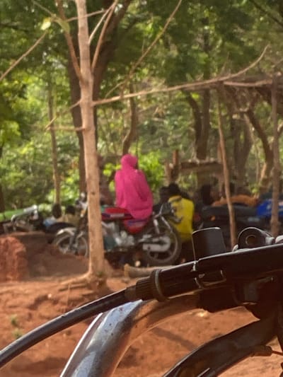 The image shows a dirt road in Egboje, Nigeria, lined with motorcycles. The foreground is dominated by the handlebars of a motorcycle, out of focus. Several motorcycles are parked along the roadside, some covered with pink and yellow cloths. Trees are visible in the background.