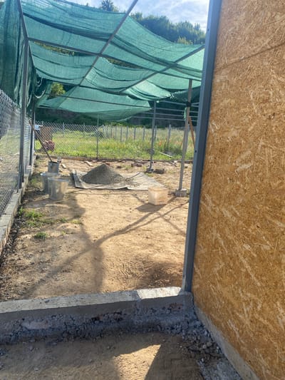 The image depicts an outdoor construction site, likely in Balgarchevo, Bulgaria, characterized by an unfinished structure and building materials. A green shade cloth is stretched over a metal frame, providing cover for the area. In the foreground, a concrete foundation runs along the edge of the site. The ground is a mixture of sand and dirt, with some scattered pebbles and grass. Several construction elements are visible, including a pile of grey gravel on a black tarp, two metal buckets, and some bricks. A fence made of wire mesh runs along the left side of the scene. To the right, a wall constructed from oriented strand board (OSB) is partially visible, revealing a metal frame support. The background shows a grassy slope with trees under a bright sky with scattered clouds. The overall impression is of an ongoing construction project in a rural or semi-rural setting.