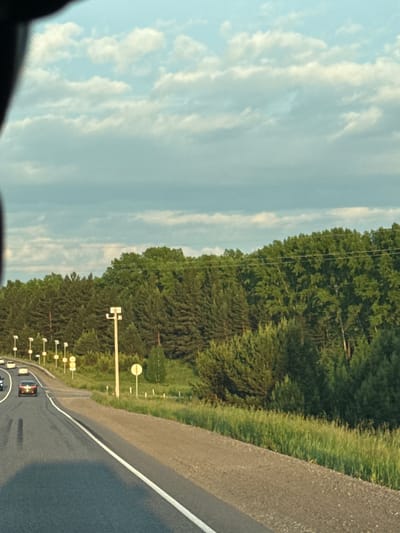 The image captures a view from a vehicle traveling on a road, likely in or near Sukhaya, Russia. The scene depicts a highway with several cars in motion. On the right side of the road, a dense forest of coniferous and deciduous trees lines the landscape, extending to the horizon. The sky above is a blend of blue with scattered, fluffy white clouds, suggesting it is daytime, possibly late afternoon due to the quality of light. Along the side of the road, there are streetlights and some road signs visible. The road surface appears to be asphalt, with a white dividing line. The overall environment suggests a rural or semi-rural setting on the outskirts of a populated area.