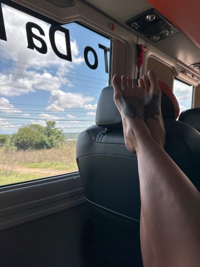 A person's feet, with tattoos on the soles, are propped up on the back of a black leather seat inside a bus. The feet are angled towards the viewer, with the right leg extending upwards and out of frame. Through the bus window, a landscape of green trees, dry grass, and a cloudy sky is visible. Black lettering on the window reads "TO DA". The scene appears to be set in Hòa Thắng, Vietnam, likely during daylight hours, as indicated by the natural lighting and the sky.