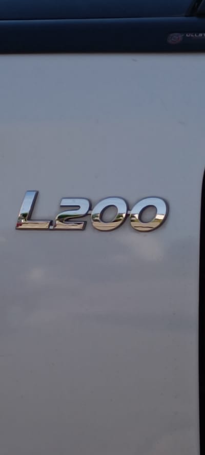 The image shows a chrome "L200" emblem on a white vehicle. The emblem is positioned horizontally, with the "L" on the left and the two "0"s on the right. The chrome reflects the surroundings, including what appears to be greenery and a blue sky. Above the emblem, a dark colored trim piece is visible, and on the far right edge of the image, a thin black vertical line suggests the edge of the vehicle. The rest of the image is a light gray, textured surface, likely the vehicle's paint. There is no indication of people, actions, or interactions. The location within Schwaikheim, Germany is not discernible from the provided image.