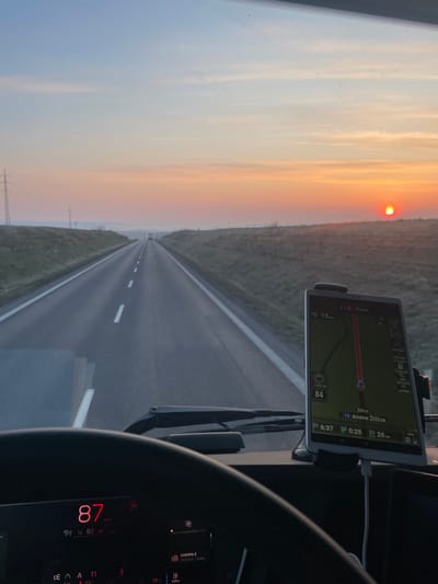 The scene depicts a view from the interior of a vehicle, likely a truck, looking forward through the windshield. The time of day is early morning, indicated by a prominent orange-red sun visible low on the horizon to the right, consistent with sunrise, and the digital clock displaying 6:37. The vehicle is traveling on a two-lane asphalt road with dashed white center lines and solid white shoulder lines, extending into a hazy distance where another vehicle is faintly visible.
On the vehicle's dashboard, a white tablet is mounted, displaying a navigation application. The tablet indicates the destination as "Praha" (Prague), 15 km away, with the vehicle's current speed at 84 (likely km/h) and a speed limit of 90. The next turn or point of interest is "16 boane Žižice". This places the vehicle in the vicinity of Slaný, Czechia, as Žižice is a village near Slaný, and the distance to Prague is consistent with this location.
The vehicle's digital instrument cluster is partially visible in the lower left, displaying "87" (indicating current speed), and "EVROPA 2" for the radio. The immediate foreground includes the top portion of a steering wheel and windshield wipers at the base of the windshield. The landscape flanking the road consists of grassy verges and fields, with utility poles or electricity pylons visible on the left side. The sky transitions from a pale blue at the top to a vibrant orange and red near the horizon due to the sunrise.
