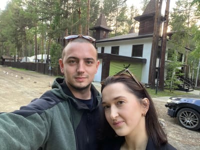 In a selfie taken outdoors in Tal'tsy, Russia, a couple stands in front of a multi-story house with distinctive turret-like roof elements. The man, on the left, has a shaved head, and sunglasses are perched on top of his head. He is wearing a dark and green zip-up hoodie. The woman, on the right, has long dark hair and is wearing a black blazer, with a necklace visible. Sunglasses rest on top of her head. Behind them, the house features white walls, dark-framed windows, and dark brown roof accents. A green banner is partially visible on the lower part of the house. The surrounding environment consists of tall pine trees and a dirt ground. To the right, the front of a dark-colored car, with a damaged bumper, is visible. Several small white posts are scattered across the ground in the mid-ground. The lighting suggests it is late afternoon or early evening.