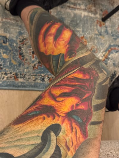 A tattooed leg is visible in the foreground, with the tattoo featuring a detailed depiction of a face, possibly a demon or mythical creature, in shades of orange, red, and blue. The tattoo extends down the leg, with abstract black elements also incorporated. The leg is resting on a rug with a blue and red patterned design. A portion of dark fabric, likely clothing, is visible at the top of the image, and a wooden floor is partially visible at the bottom. The scene is indoors. There is no information in the image to determine the location within the city of Deventer, Netherlands.
