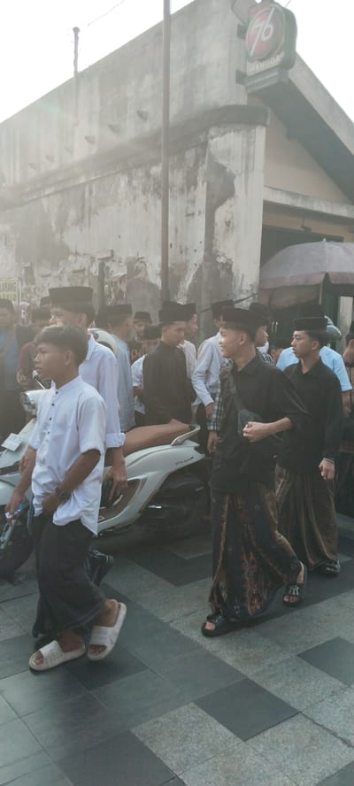 A group of young men are walking together in what appears to be an outdoor urban setting, possibly during a religious event. Most of the men are wearing traditional Indonesian clothing, including peci (caps) and sarongs. One person in the foreground is wearing a white collared shirt and black pants, while others are in black shirts and sarongs. A white scooter is parked to the left of the frame. The background shows a weathered, textured wall with some signs and an awning. The atmosphere is hazy, possibly due to sunlight or dust. The specific location is the city of Kauman, Indonesia, which is known for its rich history and Islamic heritage.