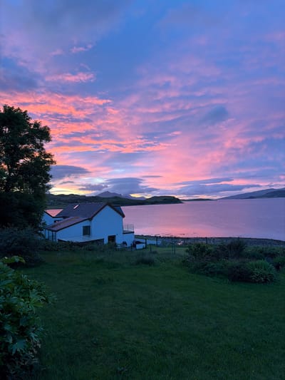 A twilight scene unfolds in Sconser, United Kingdom, featuring a modern white house with a dark roof and a glass-enclosed extension. The house is situated on a grassy slope overlooking a tranquil body of water, likely a sea loch given the surrounding hills and islands. The water reflects the vibrant colors of the sky, which is a dramatic spectacle of pink, purple, and blue clouds. A large tree with dark foliage stands to the left of the house, and a grassy lawn occupies the foreground. The shoreline is rocky, with a fence and gate visible near the water's edge. The overall impression is one of serene beauty, capturing the essence of a Scottish coastal sunset.
