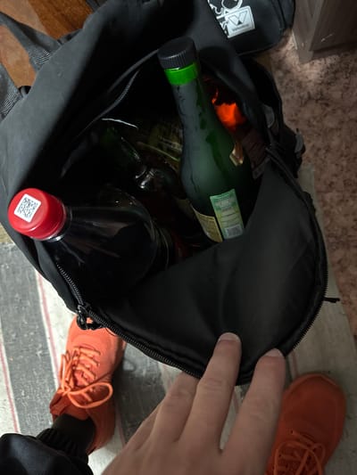 A black bag, opened by a human hand, is filled with glass bottles of various sizes and shapes, including one with a green cap. A pair of orange sneakers are visible on the floor next to the bag. The location is not discernible from the image, and there is no indication of being in the city of Маза, Russia.