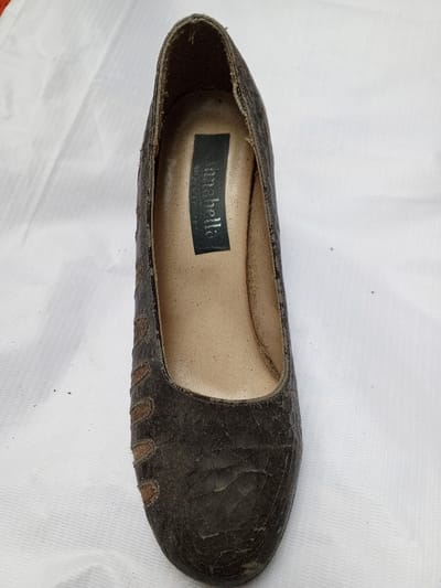 A used, dark brown, leather-like women's pump is centered in the image. The shoe has a round toe and low heel. Vertical rows of darker brown accents decorate the sides. The interior lining is a lighter tan color. A rectangular label with "Isabella" printed on it is visible inside the shoe. The background is a wrinkled white cloth. The overall condition of the shoe appears worn, with visible cracks and dirt.