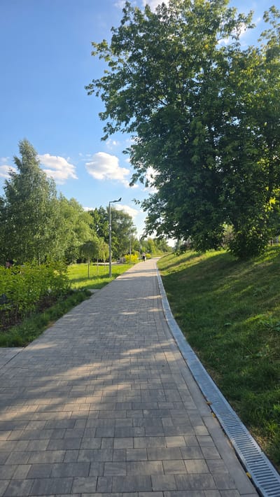 The image depicts a paved pathway in a park-like setting on a sunny day. The pathway, made of gray interlocking pavers, curves to the right, bordered by a grassy slope on its left and a metal drainage grate on its right. Sunlight casts dappled shadows across the pathway.
To the left of the pathway, lush green trees and foliage dominate the scene, with a large tree in the foreground casting shade. In the background, more trees and green grass are visible, suggesting a well-maintained park or recreational area. The sky is a clear blue with a few fluffy white clouds.
Two small figures, presumably people, are visible in the distance walking on the pathway. They are too far away to discern any details about their appearance or any specific activities they might be engaged in. The scene conveys a sense of peacefulness and a pleasant outdoor environment, likely during the daytime due to the bright sunlight and clear sky. No visible text is present in the image. The location is identified as Razvilka, Russia.