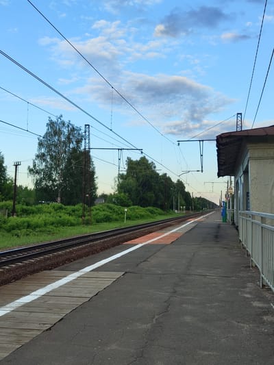 This is a ground-level, eye-level, static, medium shot of a train platform and tracks at a train station in the city of Tsibino, Russia. The train tracks run horizontally across the image from left to right, with ballast underneath. A concrete platform with a white line and an orange painted section runs alongside the tracks. A wooden ramp crosses the platform perpendicular to the tracks. To the right of the platform, a light-colored building with a brown roof and white railings is visible. Overhead, numerous power lines for the electric train are strung between tall poles and crisscross the sky. The sky is partly cloudy with a mixture of blue and white clouds. Lush green trees and bushes line the area beyond the tracks. The overall scene depicts a rural or suburban train station.