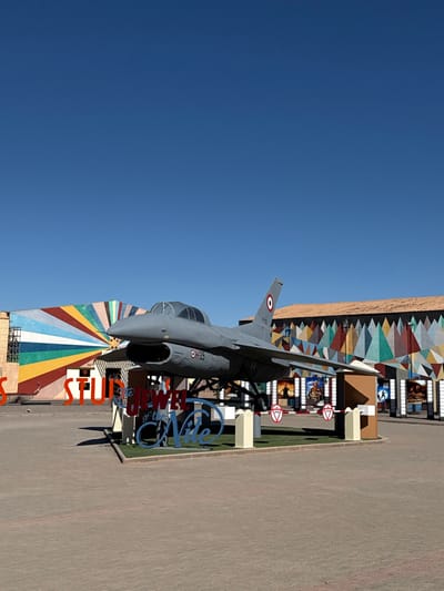 The image displays an outdoor scene at the entrance of Atlas Film Studios in Ouarzazate, Morocco, under a clear blue sky. A gray, single-engine jet aircraft, possibly a Fouga Magister, is prominently featured, mounted on a dark elevated pedestal above a patch of artificial green turf. The aircraft displays a Royal Moroccan Air Force roundel on its vertical stabilizer and the marking "OH39" on the fuselage below the cockpit. A large, stylized blue and white sign below the aircraft reads "THE JEWEL of the Nile." To the left, partially visible orange block letters spell "STUDIOS."
In the background, two distinct buildings are present. The building on the left features a vibrant mural composed of multi-colored radial stripes emanating from a central point. The building on the right has a facade decorated with a repeating pattern of multicolored geometric shapes, including triangles and squares, and is topped with a reddish-brown tiled roof. Between the aircraft and the right building, several large film posters or banners are visible, depicting various scenes.
In the foreground, the ground is paved with light reddish-brown interlocking pavers. To the right, a controlled access point is visible, featuring two beige pillars with white tops, between which are two lowered red-and-white striped boom barriers. Below each barrier, a circular red "No Entry" sign with a white horizontal bar is affixed. The scene is brightly lit by natural daylight, and no individuals or explicit actions are depicted.