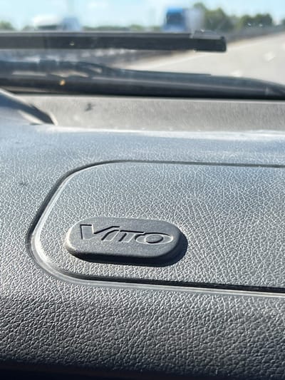 The image shows a close-up of a dark gray, textured surface, likely an interior car panel. Embossed on this surface is a logo that appears to spell "Vito". The logo is raised and has a metallic sheen, with the letters angled. To the left of the panel, out of focus, is a view through a window, showing what appears to be a road or path with trees and possibly a building in the distance. The lighting suggests it is daytime, with sunlight casting shadows on the textured surface and highlighting the embossed logo. The scene appears to be within a vehicle, possibly in a rural or suburban setting, given the blurred background. There are no people visible in the image. The primary focus is the "Vito" logo on the car's interior.