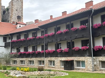 The image captures a three-story building in a serene outdoor setting, likely within the Despotovac Municipality, Serbia. The building's facade is striking, featuring a combination of stone on the ground floor and white walls on the upper floors, accented by dark wooden balconies lined with vibrant pink flower boxes. A red tile roof tops the structure, punctuated by brick chimneys. To the left of the building, a stone tower, possibly part of a historical fortification, adds a sense of age and grandeur to the location. The foreground is meticulously landscaped with a small pond, complete with a miniature waterfall, surrounded by carefully arranged rocks and flourishing vegetation, including green grass and yellow flowering plants. A small tree stands nearby. The scene is bathed in soft, overcast light, creating a tranquil atmosphere.
