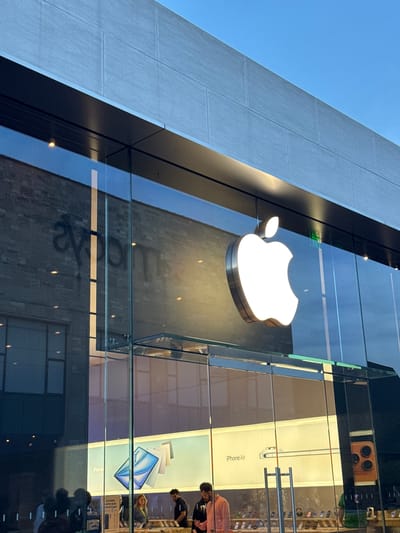 The image captures an Apple Store in Oak Brook, United States of America. The store's iconic illuminated Apple logo is prominently displayed on a glass facade. The building features a modern architectural style, with a gray stone exterior extending towards the blue sky. The store's interior, visible through the glass, reveals display cases and people browsing products. iPhone and iPad products are visible on display, as are promotional images. The reflection of the building across the street is visible on the glass.