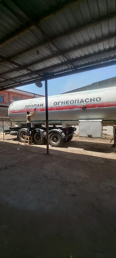 The image shows a large white tanker truck with red stripes, parked under a covered structure with a corrugated metal roof. The truck is labeled with Cyrillic text, including "ПРОПАН" and "ОГНЕОПАСНО," which translates to "PROPANE" and "FLAMMABLE." A person is visible on the side of the truck, appearing to be performing some kind of maintenance or inspection. The truck has multiple axles and large wheels. In the background, there is a brick building and a light blue sky, suggesting it is daytime. The ground is a rough, gray surface. The setting appears to be an industrial or service area, possibly a depot or maintenance yard in Almalyk, Uzbekistan. The overall impression is one of an operational vehicle in a functional environment.