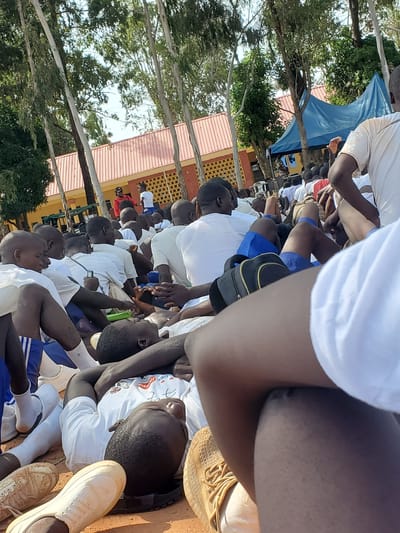 In Barakin Rafin Gora, Nigeria, a group of young men are seated on the ground, appearing to be gathered outdoors near a building with a red corrugated metal roof and yellow and orange patterned walls. Tall, slender trees with green foliage form a backdrop, and a blue tent is visible in the background. Most of the individuals are wearing white t-shirts and shorts, with some wearing white socks and athletic shoes. The perspective is close-up, with some individuals in the foreground and others further back. One person is lying down on their back, looking upwards. Several people are holding or interacting with cell phones. The overall impression is a casual gathering or assembly.