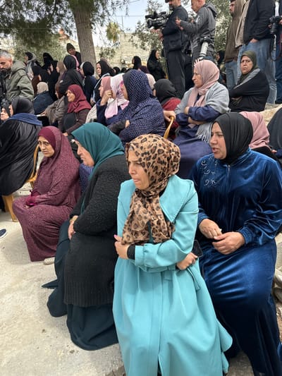 The image depicts an outdoor gathering in Dura, Palestinian Territory. The foreground and midground are densely populated by numerous individuals, predominantly women, many of whom are seated. Women are dressed in long, modest clothing and head coverings (hijabs) of various colors, including burgundy, dark teal, black, light blue, grey, and a leopard print pattern. In the immediate foreground, a woman wearing a light blue coat and a leopard print hijab shows an expression consistent with distress or crying, with tears visible on her face; she holds a dark cell phone in her left hand. To her right, another woman in a dark blue velvet abaya and black hijab is speaking, with her mouth open. In the background, several men are standing, some operating professional video cameras with attached lenses and possibly tripods, indicating a media presence. The ground is a light-colored concrete surface. Trees and distant building structures are visible under an overcast sky. Yellow-gold chairs are partially visible where some women are seated. The overall atmosphere appears somber.