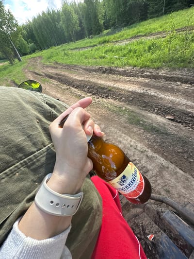 A first-person perspective image shows a person seated outdoors, holding a beer bottle labeled "Hoegaarden Grapefruit." The person is wearing green pants, a white sweater, and a white sports watch. The legs of the person are covered by a red blanket. The background features a dirt road alongside a grassy area with trees. The location is potentially Bogorodskoe, Russia.