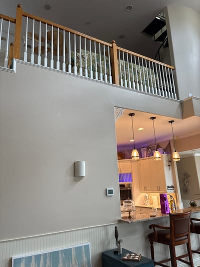 The image shows an interior view of a residential dwelling, likely a home, with a multi-level design. In the foreground, a white beadboard wainscoting runs horizontally along the lower portion of the wall. Above this, a large, smooth, beige-painted wall dominates the left and center of the frame. On this wall, a white cylindrical light fixture is mounted.
The upper portion of the image reveals a mezzanine or upper floor with a white wooden railing. Behind the railing, a ceiling fan with large, light-colored blades is visible, and patterned upholstery, possibly on a sofa or cushions, is glimpsed. Further in the background, a spiral staircase with black metal steps is visible, suggesting a second level or loft area.
To the right of the main beige wall, an opening reveals a kitchen area. Three pendant lights with glass shades hang above a granite-topped counter. The kitchen features light-colored cabinets, a microwave or oven, and some decorative items on the counter, including a purple-wrapped object, a bottle, and a glass. A seahorse decoration is mounted on the wall behind the counter.
The overall impression is of a modern, open-plan living space with a coastal or tropical aesthetic, suggested by the seahorse and possibly the upholstery pattern.
This scene is located in Bradenton, United States.