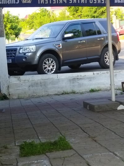 A gray Land Rover Freelander SUV is parked on a sidewalk in front of a building with a blue awning that has white text on it. The car is facing towards the left side of the image and is partially obscured by a white pillar. The building appears to be a business as indicated by the awning. The sidewalk is paved with gray square tiles, and some patches of grass are growing through the cracks. The background shows green trees and a road. There is no indication of any people in the image.