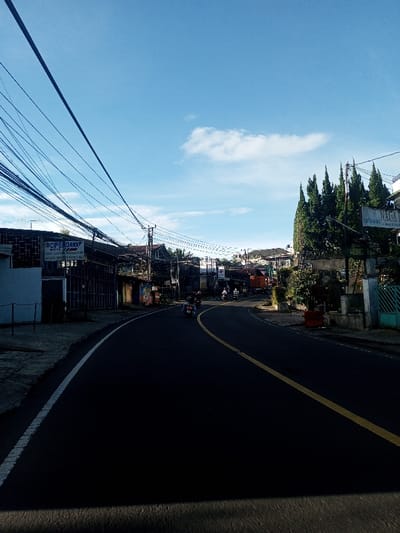 The image presents an urban street scene in Khouathineung, Laos, viewed from a low angle on the asphalt road under a bright blue sky with sparse white clouds. The paved road curves gently to the right, delineated by a continuous white line along the left shoulder and a continuous yellow line near the center. Numerous dark utility wires span overhead across the left side of the frame.
On the left side of the road, a series of multi-story buildings line the street, appearing to be commercial establishments. One building prominently features a sign with text including "CIPE DAWA MOTOR." A sidewalk or unpaved path runs alongside these structures.
On the right side of the road, additional buildings are interspersed with mature vegetation, including several tall, slender conifer-like trees. A white sign with dark lettering, partially obscured, is visible amongst the trees, identifying "NAGA RESTAURANT." Another sign below it appears to read "HONG LY." Further down, a light-colored building with a distinctive blue-green gate is visible.
Several individuals on motorcycles are present on the road, traveling away from the viewer towards the curve. One motorcycle is visible in the mid-ground, and multiple others are further ahead. A large orange truck is also observed further down the road on the right side. Shadows are cast across the road surface, suggesting direct sunlight.