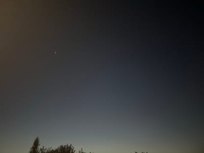 The image captures a dark, dusky sky above a silhouetted line of trees. Two distinct celestial bodies are visible in the sky. One appears as a bright, elongated smear with multiple points of light, while the other is a smaller, single point of light. The sky transitions from a yellowish-orange glow on the left to a deep blue-grey on the right. The trees at the bottom of the frame are dark and indistinct, with the tops of several pine trees and other foliage visible. The scene is consistent with a nighttime or twilight photograph taken in an outdoor setting, likely in or near Lyubuchany, Russia, given the provided context. There are no people or discernible human activity depicted in the image.