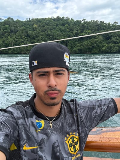A close-up selfie captures a young adult male on a boat in the coastal waters of Mangaratiba, Brazil. The male, positioned centrally and facing forward, has a medium complexion, short dark hair, brown eyes, a mustache, and a light goatee. He wears a black fitted baseball cap featuring an MLB logo on the front left panel and a colorful "Los Angeles Dodgers" script logo on the right side panel. His facial piercings include a small silver stud below the outer corner of each eye and a silver hoop in the right nostril. He is dressed in a short-sleeved grey and black athletic-style t-shirt adorned with a graphic featuring a depiction of Jesus Christ, a yellow Nike swoosh, a CBF (Brazilian Football Confederation) shield with five stars, and a yellow and blue emblem resembling the Brazilian flag's circle. A thin gold chain with a small gold cross pendant is visible around his neck. The background consists of calm, grey-green water with ripples, a densely forested landmass or island in the distance, and a partly cloudy sky. A varnished wooden boat railing, showing water droplets, is visible at the lower right edge of the frame.